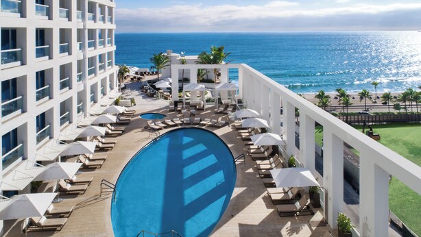 A hotel pool area with a view of the ocean, lounge chairs, umbrellas, and a large swimming pool. - Luxury Suites at Fort Lauderdale Beach (Fort Lauderdale)