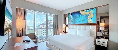 Une chambre d’hôtel avec un grand lit, un bureau, une chaise et une grande fenêtre donnant sur des immeubles et la mer.