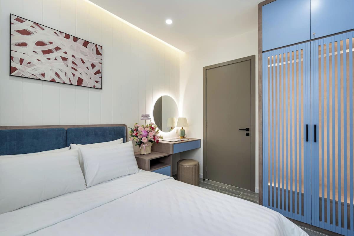 A modern hotel room with a bed, a desk with a lamp, a mirror, and a wardrobe.