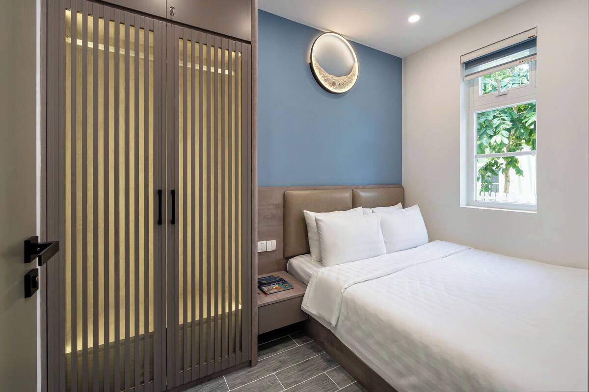 A bedroom with a bed, a nightstand, a wardrobe, a mirror, and a window with a view of greenery.