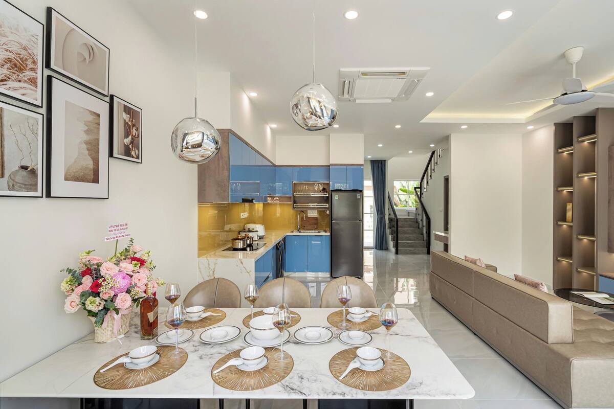 A modern dining area with a marble table set for six, a kitchen in the background, and a staircase leading to another level.
