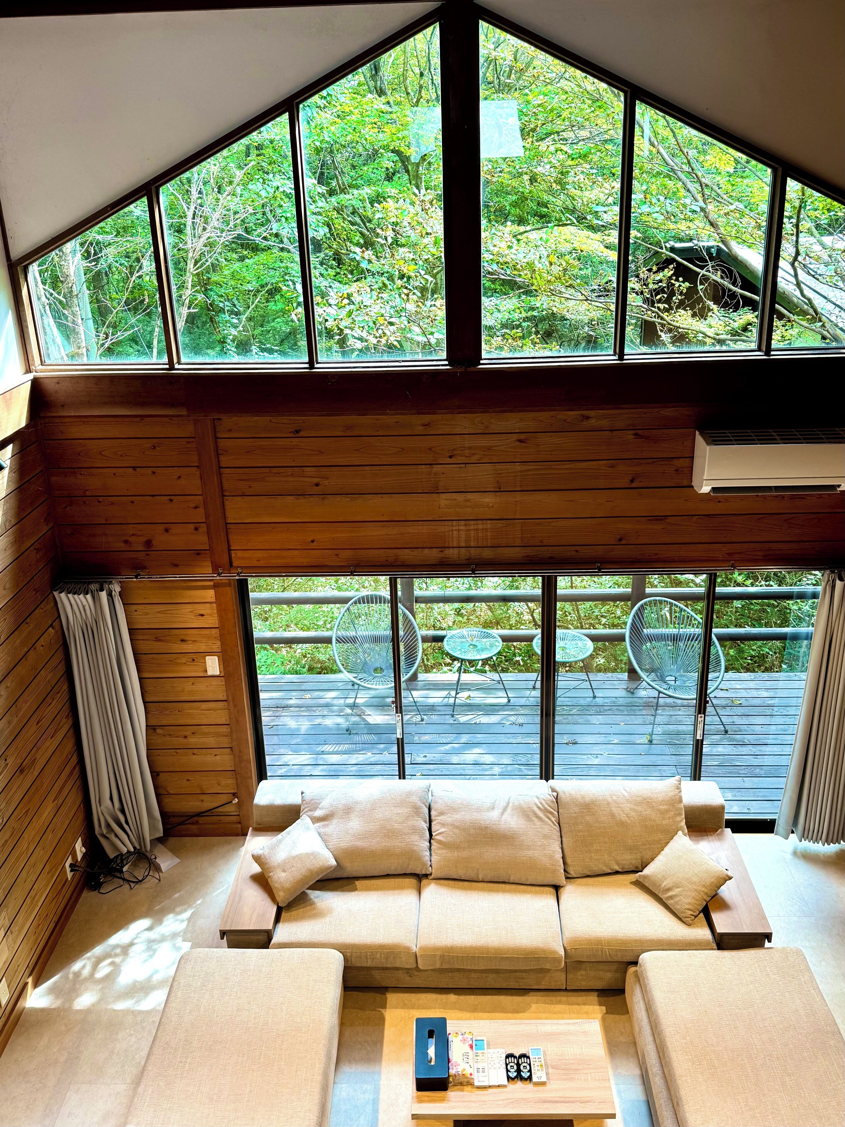 A cozy room with a wooden ceiling, a beige sofa, and a small table with remote controls. Large windows offer a view of greenery outside.