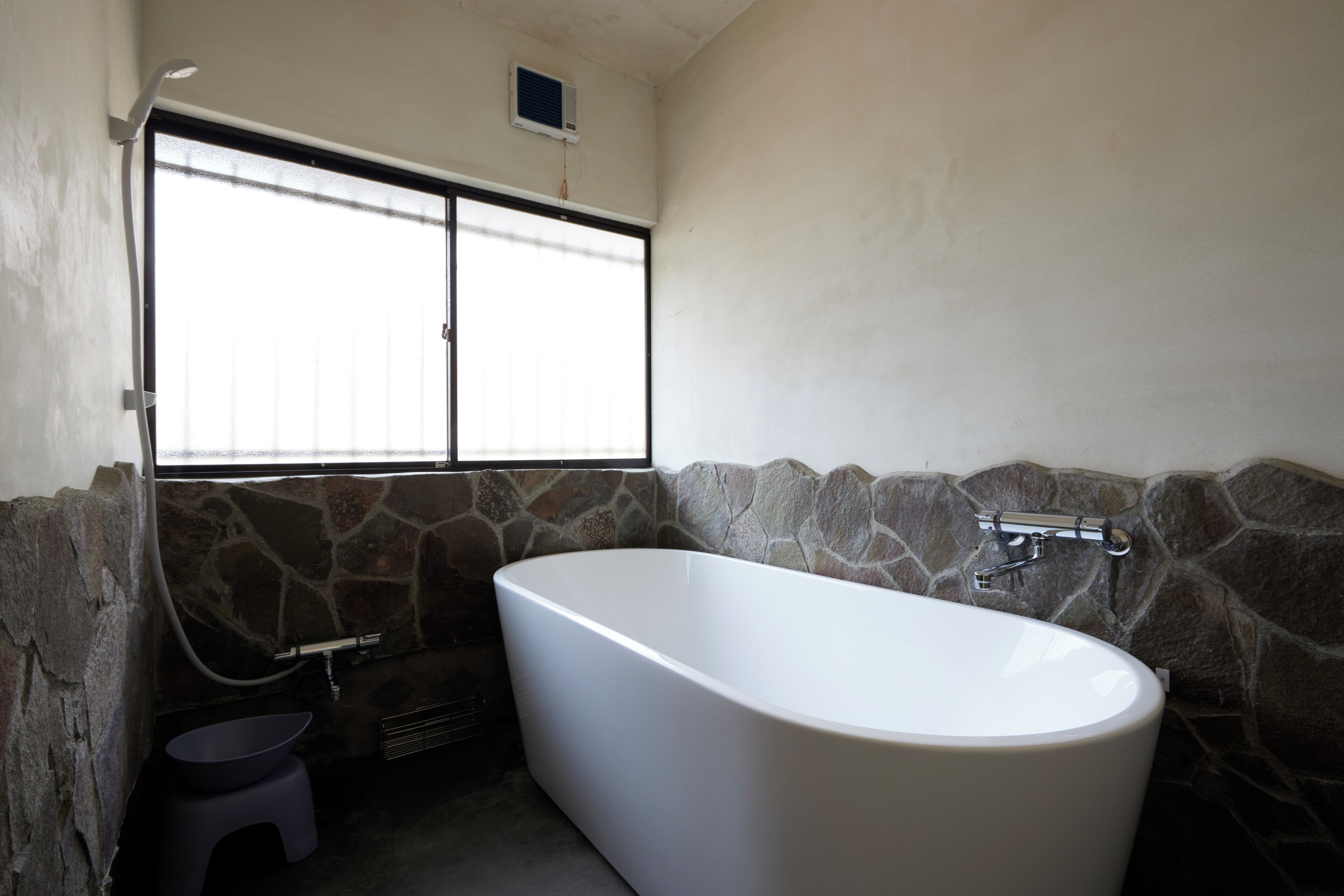 A bathroom with a freestanding bathtub, a window with blinds, and stone wall accents.
