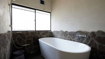 A bathroom with a freestanding bathtub, a window with blinds, and stone wall accents.