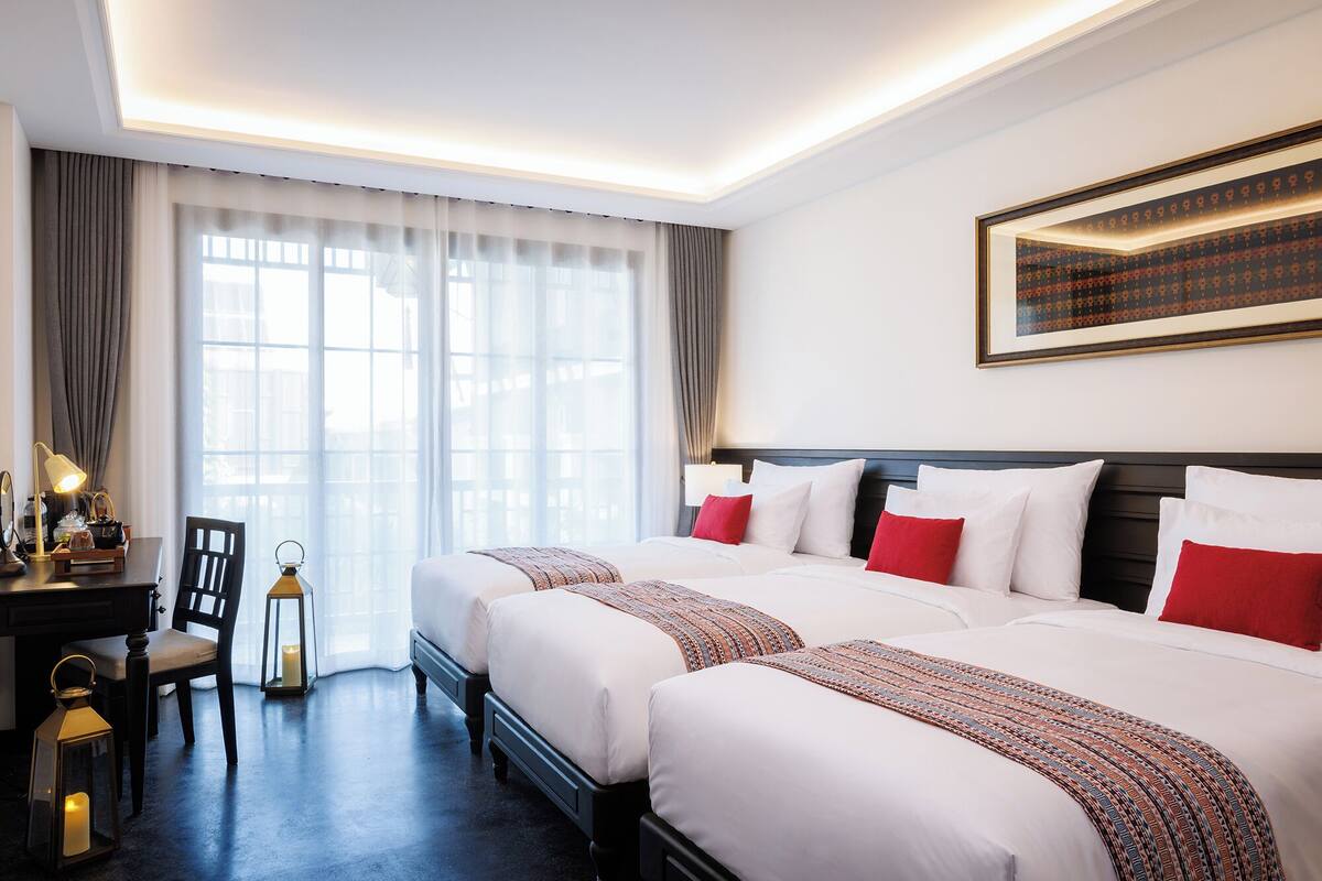 A hotel room with two beds, a desk, a chair, a table with a lamp, and a window with curtains.
