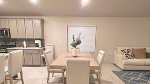 A modern living room with a dining area, featuring a wooden dining table, beige chairs, a sofa, and a coffee machine. - The paradise house in Roswell (Roswell)