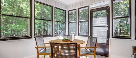 A room with a round table and four wicker chairs, large windows with a view of trees, and a door with a window.
