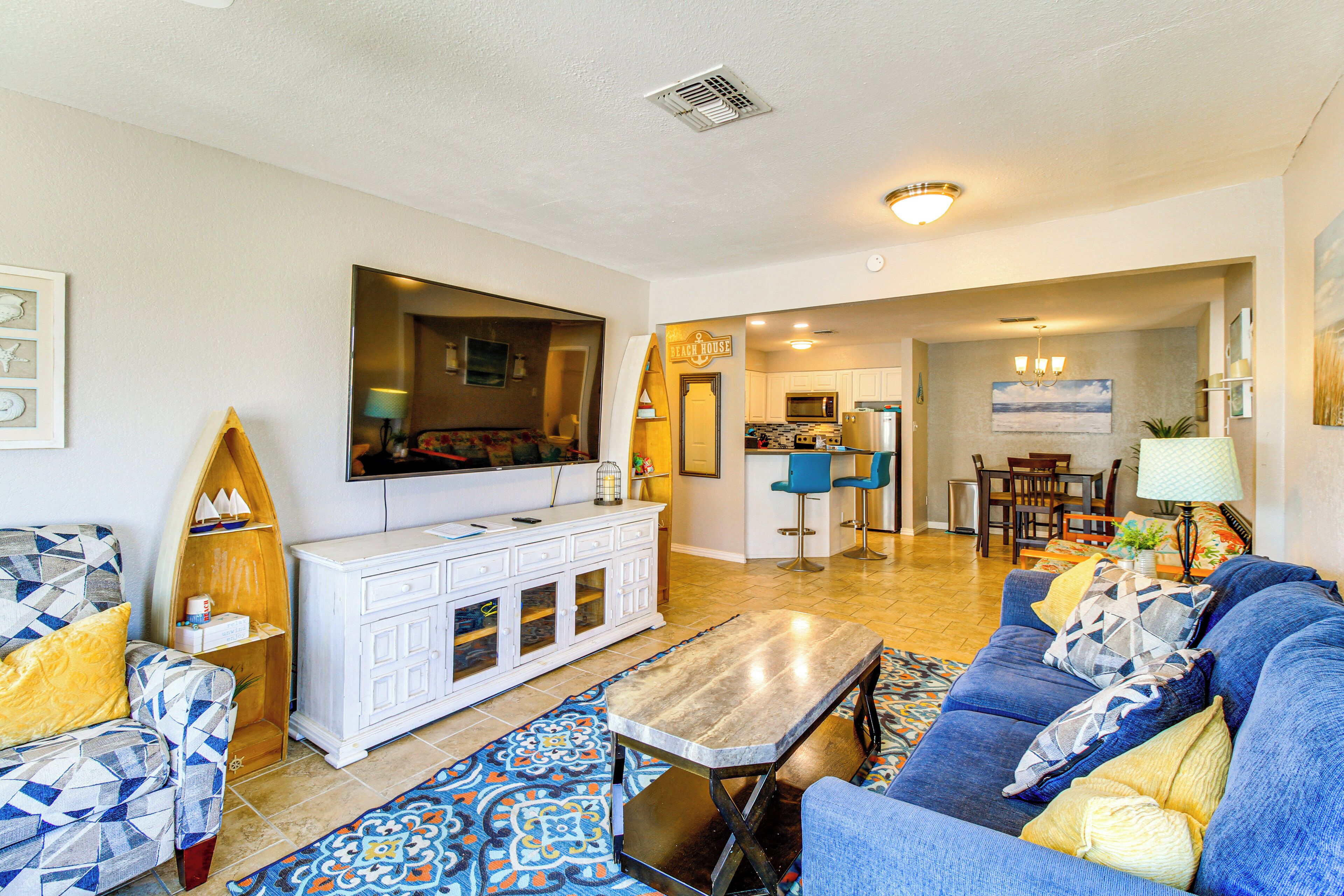 A living room with a sofa, coffee table, and a television. There is a kitchen area in the background with a dining table and chairs.
