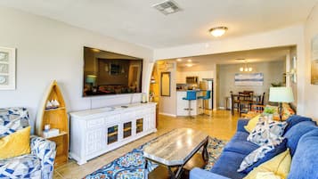 A living room with a sofa, coffee table, and a television. There is a kitchen area in the background with a dining table and chairs.