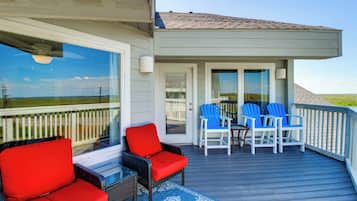 A covered outdoor deck with wicker chairs and a table, a glass door leading to a covered porch, and a view of the landscape.
