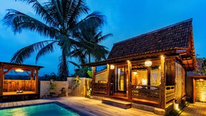 A wooden house with a pool, surrounded by palm trees, at dusk. - Kamania Villa Ubud by Dhananjaya Hospitality (Sebatu)