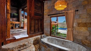 A bathroom with a stone wall, a bathtub, a wooden door, and a window with curtains. - Kamania Villa Ubud by Dhananjaya Hospitality (Sebatu)
