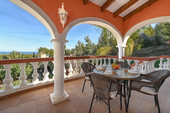 A covered patio with a table set for four, surrounded by wicker chairs, overlooking a scenic landscape with trees and a view of the sea.