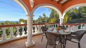 Un patio cubierto con una mesa preparada para cuatro, rodeado de sillas de mimbre, con vistas a un paisaje pintoresco con árboles y el mar.