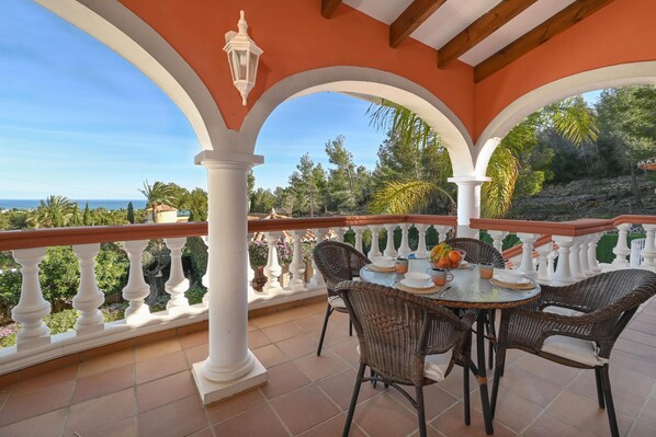 Une terrasse extérieure couverte, avec une table ronde et des chaises en osier, entourée de balustrades blanches et offrant une vue sur l’océan.