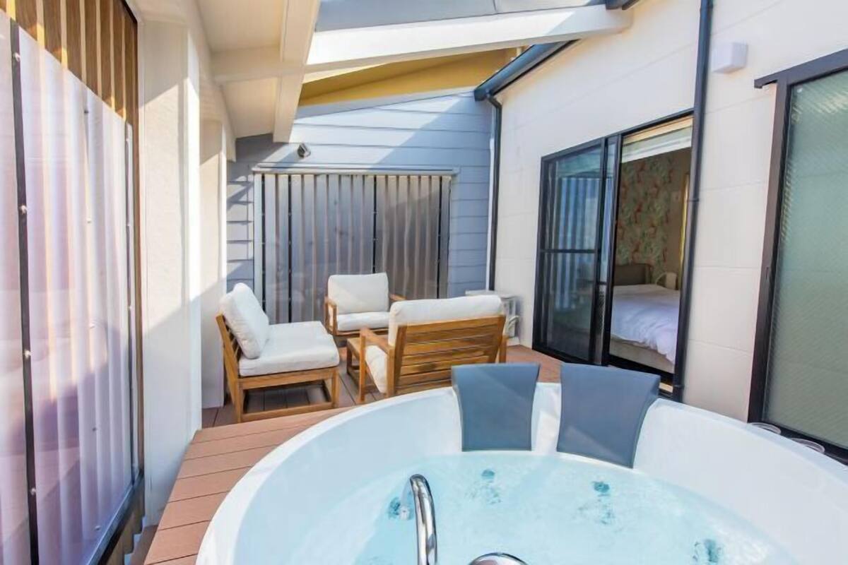 A sunlit outdoor area with a hot tub, wooden chairs, and a bed with a window.
