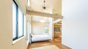 Une chambre avec un lit, une fenêtre, un ventilateur de plafond et, au fond, un coin cuisine.