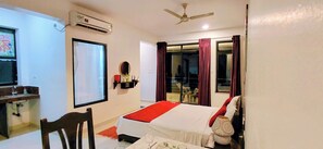 A hotel room with a bed, a chair, a table, a mirror, a window, and a ceiling fan. - Wave Morjim Hotel (Morjim)