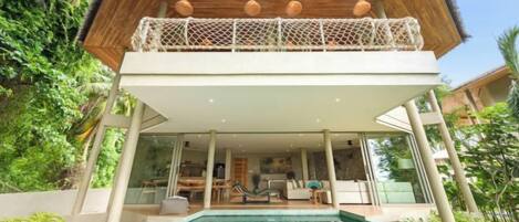 A modern villa with a glass-enclosed pool, a wooden deck, and a thatched-roof upper level.