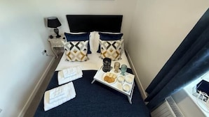 Ein ordentlich gemachtes Bett mit gemusterten Kissen, ein Nachttisch mit Lampe und eine Ablage mit Speisen und Getränken.