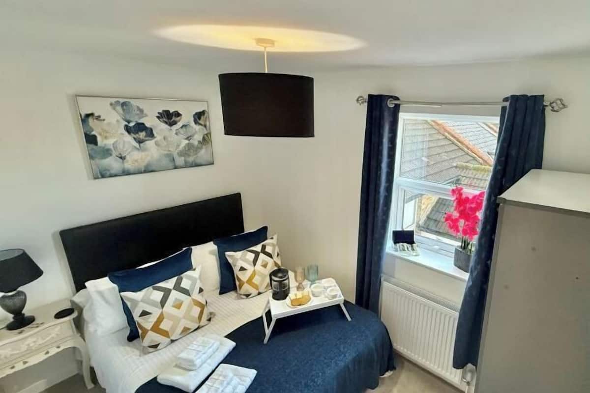 A bedroom with a bed, bedside table, lamp, artwork, window with curtains, and a view of the outside.