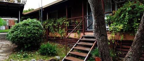 A traditional building with a wooden staircase leading to a covered porch, surrounded by greenery and a stone pathway.