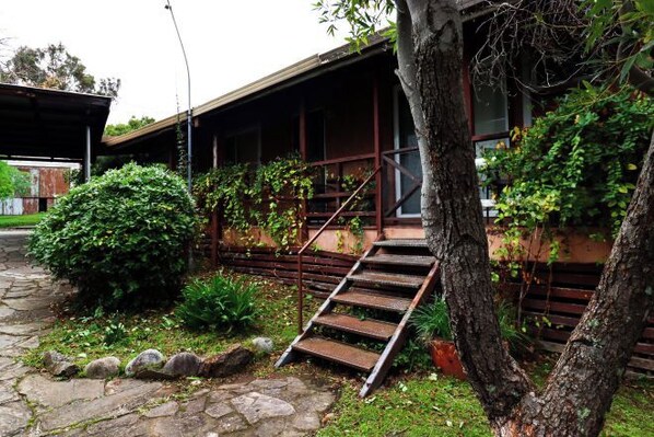 A traditional building with a wooden staircase leading to a covered porch, surrounded by greenery and a stone pathway. - Nature Lovers: Snowy Cottage Escape! Book Direct (Talbingo)