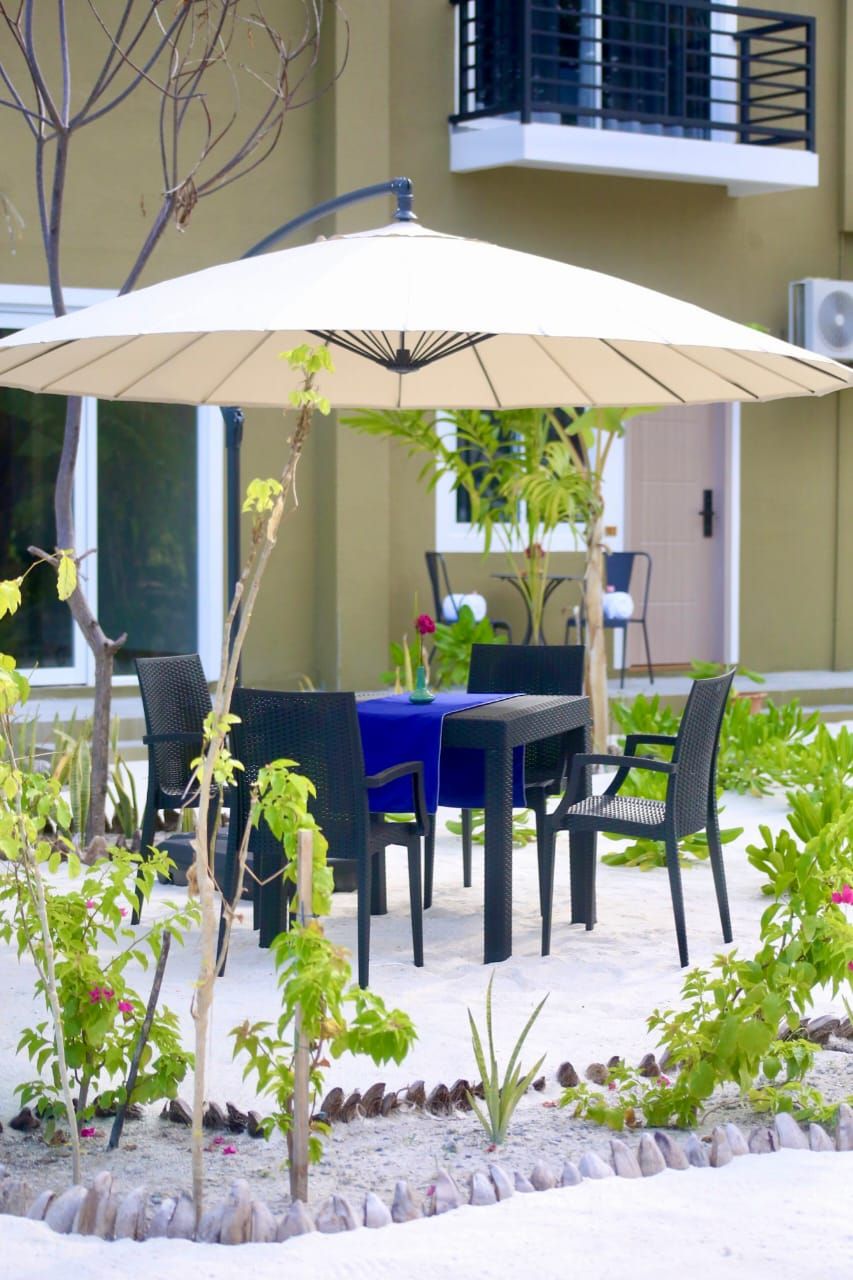 An outdoor seating area with a table and chairs under a large umbrella, surrounded by plants and a stone border.