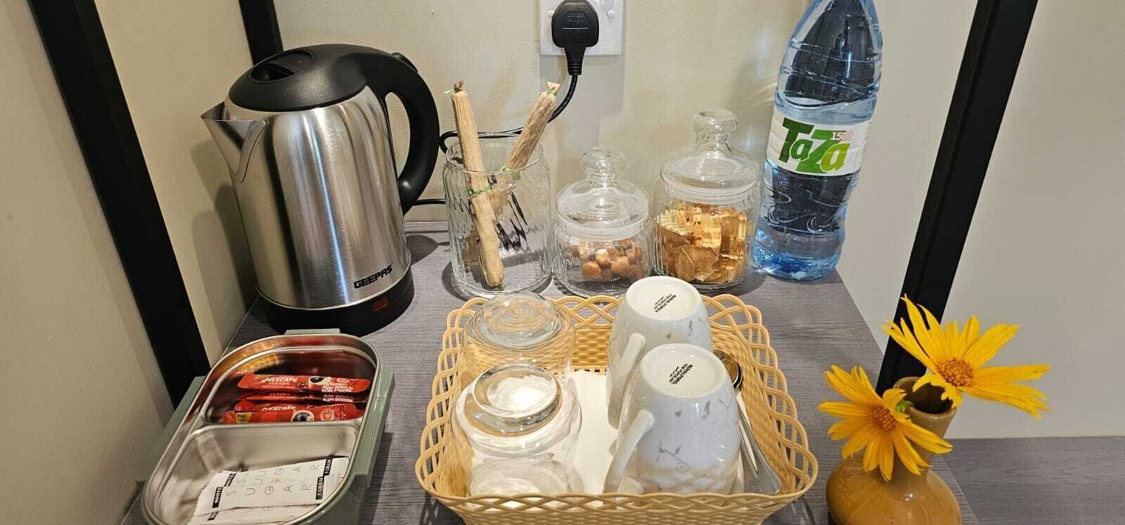 Una encimera de cocina con un hervidor, frascos, una cesta con tazas, una botella de agua y un florero.