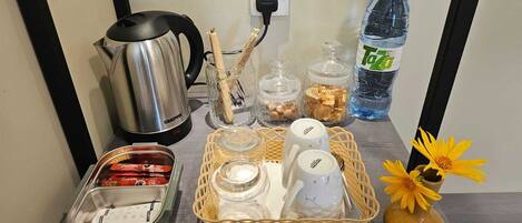 Una encimera de cocina con un hervidor, frascos, una cesta con tazas, una botella de agua y un florero.