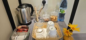 Un comptoir de cuisine avec une bouilloire, des bocaux, un panier rempli de tasses, une bouteille d’eau et un vase à fleurs.