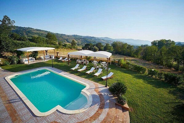 A swimming pool with lounge chairs and umbrellas, surrounded by a landscaped garden and hills in the background.