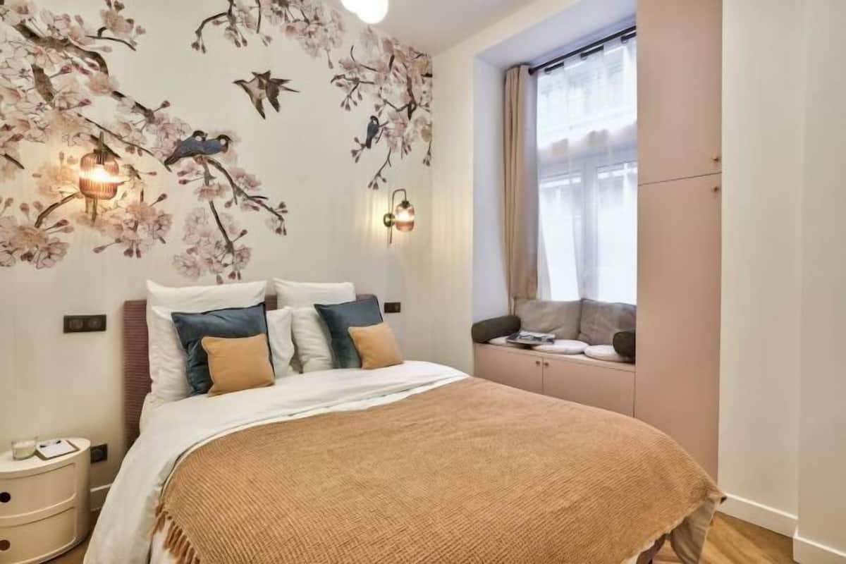 A bedroom with a bed, pillows, a nightstand, a window with curtains, and wall-mounted lights.