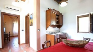 Une pièce avec un sol carrelé, une table à manger recouverte d’une nappe rouge, un panier en osier, des meubles en bois et une fenêtre avec des rideaux.