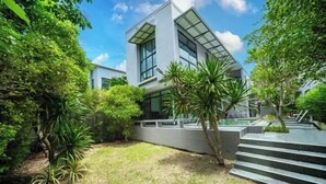 A modern building with large windows and a flat roof, surrounded by lush greenery and a paved area with steps leading up to the entrance.