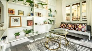 Een woonkamer met een bank, twee salontafels met glazen blad en een boekenkast vol boeken en decoratieve voorwerpen.