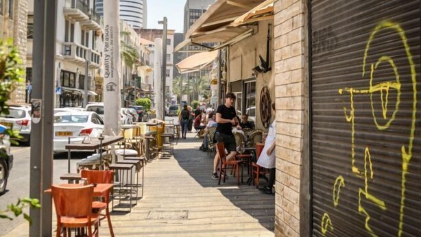 An outdoor café with tables and chairs, a closed shutter on the right, and a street with cars and buildings in the background. - The Printhouse Boutique Studio 4 (Haifa)