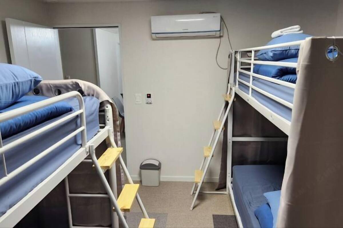 A dormitory room with bunk beds, a white air conditioning unit, a door, and a trash bin.