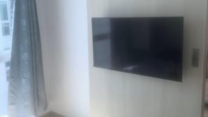 Una habitación con televisión de pantalla plana en la pared, una ventana con cortina y pared de color claro.