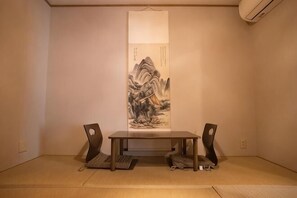 Um quarto com piso de esteira de tatame, uma mesa baixa com duas cadeiras e um pergaminho tradicional japonês na parede.