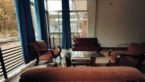 Ένα δωμάτιο με ένα τραπέζι από γυαλί και τέσσερις καφέ καρέκλες, ένα μεγάλο παράθυρο με μπλε κουρτίνες και θέα σε ένα κτίριο που βρίσκεται έξω.