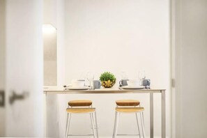 A clean, minimalist dining area with a white table set for two, a small plant, and a kettle.