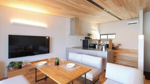A modern living room with a wooden dining table, white sofas, a flat-screen TV, and a kitchen area with a bench. - Dannoura Strait Villa (Shimonoseki)