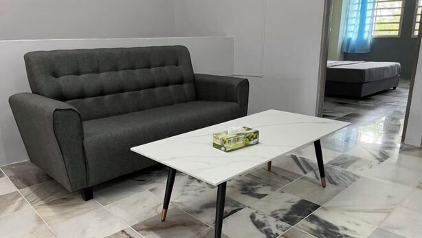 Un sofá gris y una mesa de centro blanca con una caja de pañuelos encima, en una habitación con suelo de baldosas grises y una cama al fondo.
