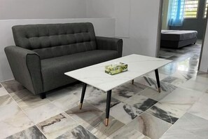 Ein graues Sofa und ein weißer Couchtisch mit einer Taschentuchbox darauf, in einem Raum mit grau gefliestem Boden und einem Bett im Hintergrund.