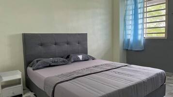 A bed with a grey headboard and pillows, a bedside table, and a window with blue curtains.