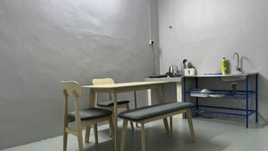 Una habitación con una mesa de madera, dos sillas, un banco, un lavamanos con una estantería azul y un hervidor.