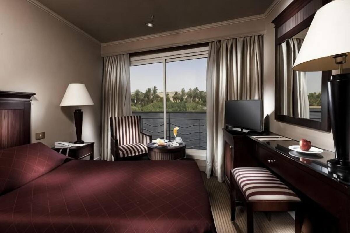 Ein Hotelzimmer mit einem großen Bett, einem Fernseher, einem Schreibtisch mit Lampe, einem Sessel und Blick ins Freie.