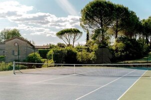 Cancha de tenis con red, rodeada de árboles y un edificio de piedra.