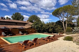Una terraza de madera con piscina, sillas de descanso y un edificio rústico con tejado de baldosas.